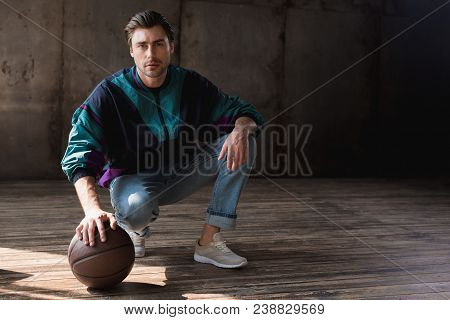 Attractive Young Man In Vintage Windcheater With Basketball Ball Sitting Squats On Wooden Floor