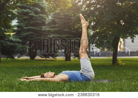 Young Man Lying Down On Green Grass Doing Lower Abs Exercise By Lifting His Legs Up. Sporty Guy Work