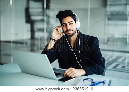 Indian Young Businessman In Earphones With Laptop Computer Working At Office And Listening To Music