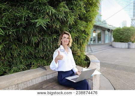 Successful Businesswoman Working With Laptop And Papers In Open Air Near Green Plant, Showing Thumbs