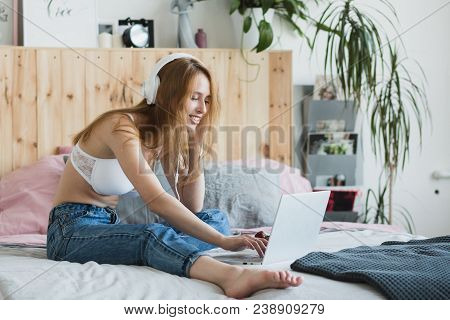 Young Sexy Blonde Woman Flirting Online On Laptop, Sitting On Bed In Bedroom. She Wearing Bra, Jeans