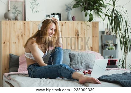 Young Sexy Blonde Woman Flirting Online On Laptop, Sitting On Bed In Bedroom. She Wearing Bra, Jeans