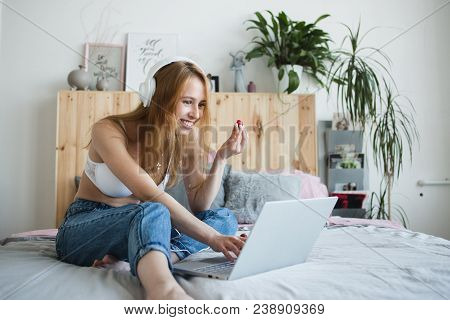 Young Sexy Blonde Woman Flirting Online On Laptop, Sitting On Bed In Bedroom. She Wearing Bra, Jeans