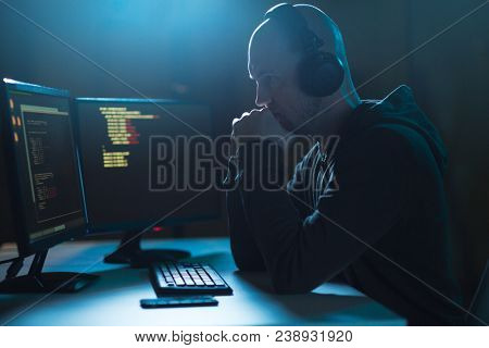 cybercrime, hacking and technology concept - male hacker with headphones and coding on laptop computer screen wiretapping or using computer virus program for cyber attack in dark room