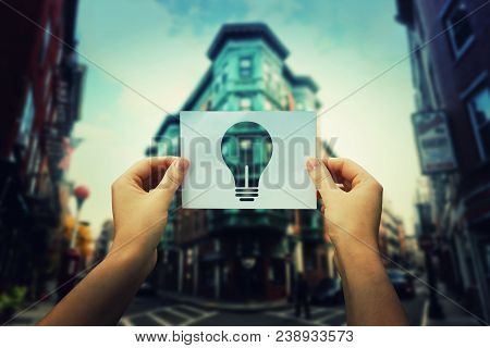 Woman Hands Holding A Paper Sheet With Bulb Icon In The Middle Of The Street In A Big City. Finding 