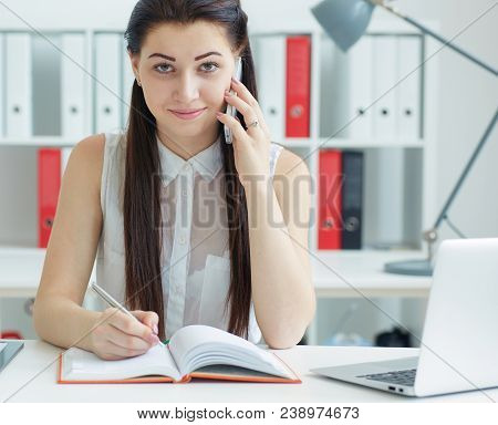 Beautiful Smiling Young Secretary Talking On The Mobile Phone Sitting At Office. Business, Exchange 