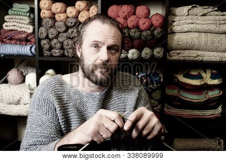 European Man 40-50 Years Old Knits A Scarf. A Man In A Knitted Sweater On The Background Of Shelves 