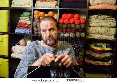 European Man 40-50 Years Old Knits A Scarf. A Man In A Knitted Sweater On The Background Of Shelves 