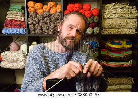 European Man 40-50 Years Old Knits A Scarf. A Man In A Knitted Sweater On The Background Of Shelves 