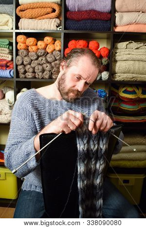 European Man 40-50 Years Old Knits A Scarf. A Man In A Knitted Sweater On The Background Of Shelves 