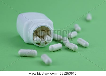 A Group Of White Capsules Of Medicine Spilled Out Of A White Jar On A Green Background. Horizontal, 