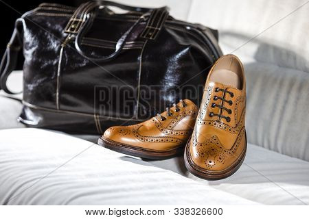Closeup Of Pair Of Male Full Brogued Tan Oxford Shoes Placed With Leather Men's Bag On Pale Sofa Ind