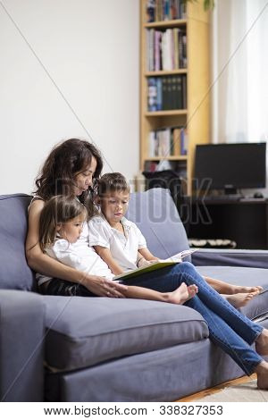 Family Ideas And Concepts. Mother Reading A Book Together With Her Little Children On Couch Indoors.