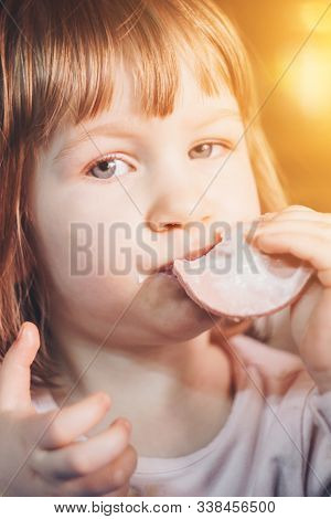A One-year-old Smiling Girl Sits At A Children's Table In A High Chair And Eats With A Spoon From A 