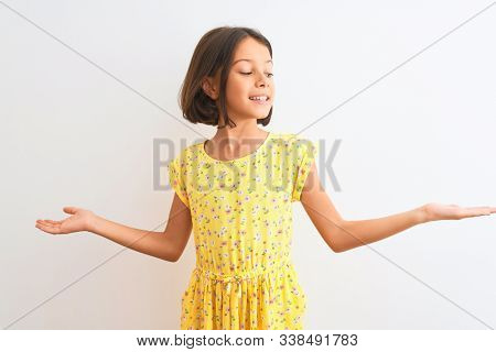 Young beautiful child girl wearing yellow floral dress standing over isolated white background smiling showing both hands open palms, presenting and advertising comparison and balance