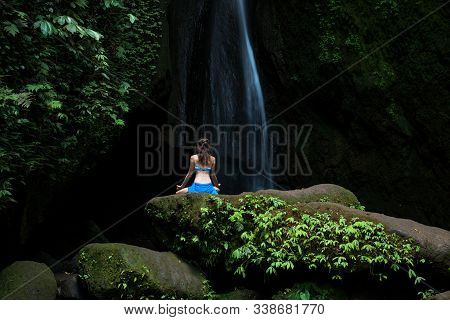 Young Woman Sitting On The Rock, Practicing Yoga Near Waterfall. Hands In Gyan Mudra. Leke Leke Wate