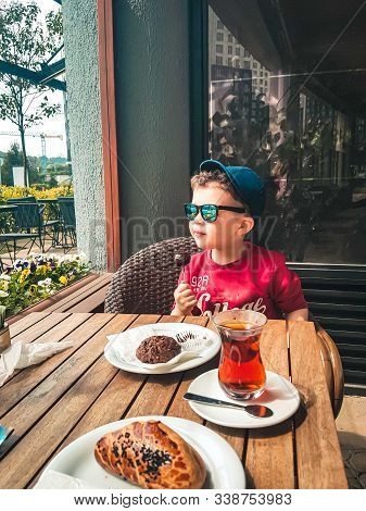 Cute Little Boy In Trendy Sunglasses And A Hat Eating Sweets At A Wooden Table In A Outdoor Cafe. Af