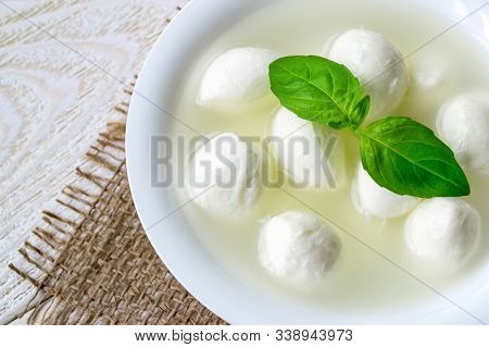 Close-up Of Bowl With Mini Mozzarella Cheese Balls In A Brine And Basil On A  White Rustic Wooden Ta