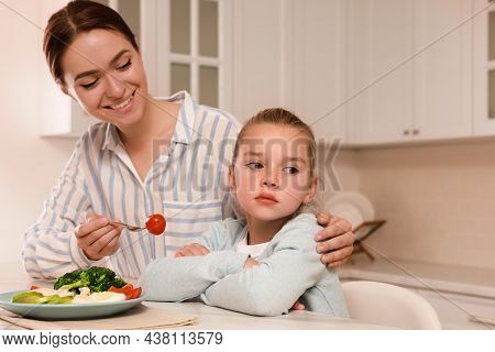 Mother Feeding Her Daughter In Kitchen. Little Girl Refusing To Eat Dinner