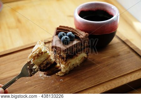 Piece Of Kiev Cake With Chocolate Icing And Blueberries And Cup Of Coffee On Wooden Board. A Bite Of