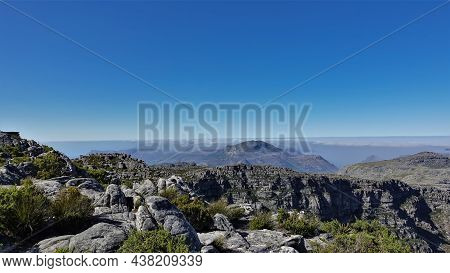 At The Top Of Table Mountain In Cape Town There Is Scant Vegetation, Gray Boulders. Clear Blue Sky. 