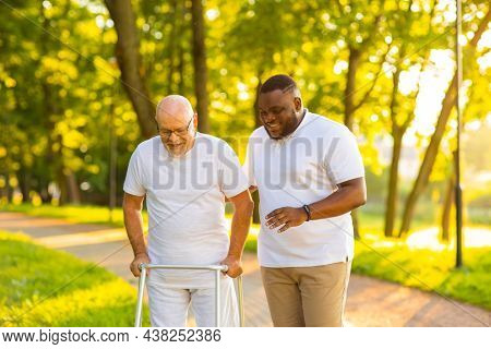Caregiver is teaching old man to walk with walker. Professional nurse and patient walking outdoor at sunset. Assistance, rehabilitation and health care.