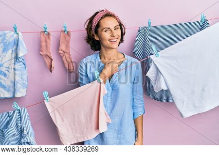 Young brunette woman doing laundry around string hangs cheerful with a smile of face pointing with hand and finger up to the side with happy and natural expression on face 