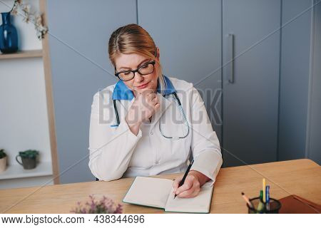 Middle-aged Nurse In White Clothes Is Sitting At A Desk In Hospital And Writing Something In A Noteb