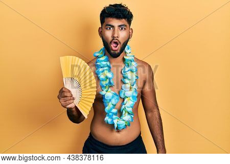Arab man with beard wearing hawaiian lei holding handfan scared and amazed with open mouth for surprise, disbelief face 