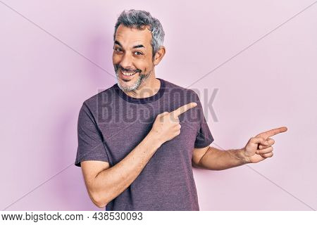 Handsome middle age man with grey hair wearing casual t shirt smiling and looking at the camera pointing with two hands and fingers to the side. 