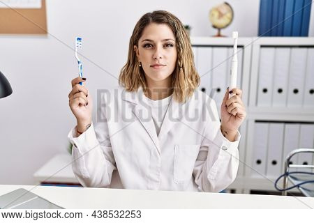 Young hispanic dentist woman holding electric toothbrush and teethbrush at clinic relaxed with serious expression on face. simple and natural looking at the camera. 