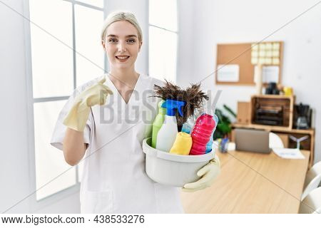Young caucasian woman wearing cleaner uniform holding cleaning products cleaning office pointing finger to one self smiling happy and proud 