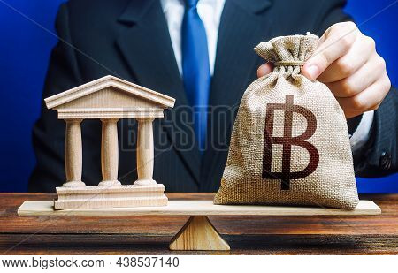 Man Puts A Thai Baht Money Bag On Scales Next To A Bank. Taxes Payment. Deposits, Grant Loans. Lobby