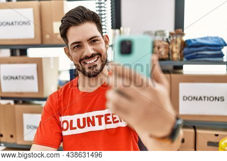 Young hispanic volunteer man making selfie by the smartphone working at charity center.