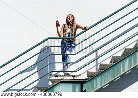 Black Woman With Coloured Braids, Looking At Her Smartphone With Her Feet Resting On A Skateboard.