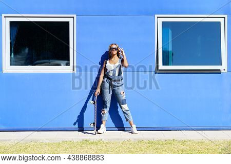 Black Woman Dressed Casual, With Modern Sunglasses And A Skateboard On Blue Wall Background.
