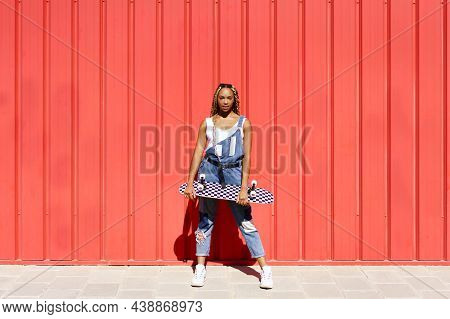 Black Woman Dressed Casual, Wtih A Skateboard On Red Urban Wall Background.