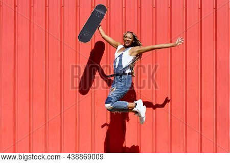 Black Woman Dressed Casual, Wtih A Skateboard Jumping With Happiness On Red Urban Wall Background.