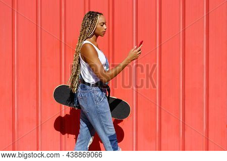 Black Woman With Coloured Braids, Consulting Her Smartphone With Her Feet Resting On A Skateboard.