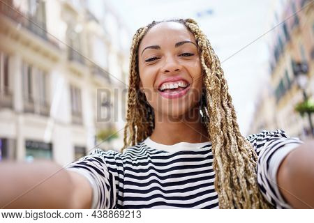 Black Woman With Afro Braids Taking A Smiling Selfie In An Urban Street With A Smartphone.