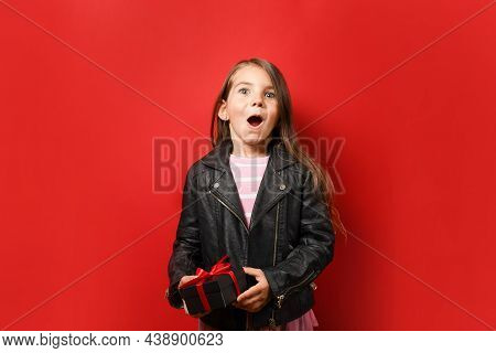 Surprised Child Girl Holding Black Gift Box On Red Background With Copy Space. Black Friday Concept