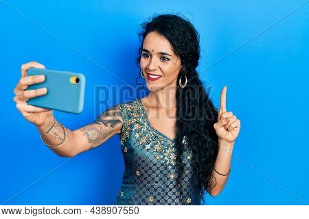 Young woman wearing bindi and traditional kurta dress taking a selfie photo with smartphone smiling with an idea or question pointing finger with happy face, number one 