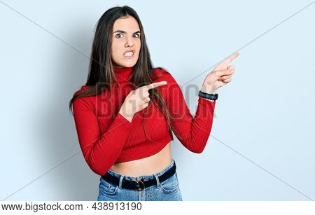 Young brunette teenager wearing red turtleneck sweater pointing aside worried and nervous with both hands, concerned and surprised expression 