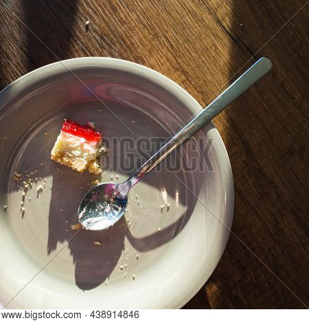 Last bite of strawberry jam cheesecake and used spoon on white plate on wooden table in morning sunlight, Half eaten cake unhealthy sweet food