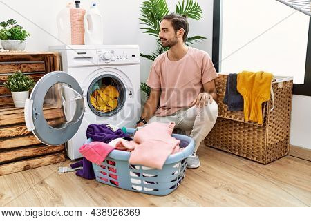 Young hispanic man putting dirty laundry into washing machine looking to side, relax profile pose with natural face with confident smile. 
