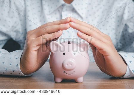 Piggy Bank Under Hands In Gesture Of Protection On Wooden Desk, Saving, Charity,  Fundrasing Communi