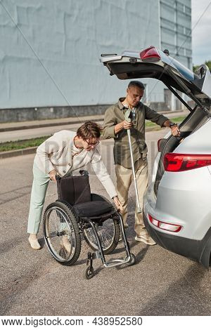 Full Length Portrait Of Mature Couple Loading Wheelchair To Car Trunk In Parking Lot Outdoors
