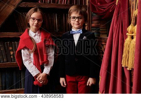 Pair of schoolchildren posing in a splended vintage interior by a bookcase in stylish, fashionable school clothes. Kid's school fashion. 