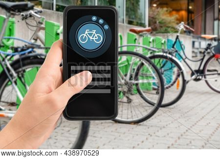 technology, transport and sustainability concept - close up of hand holding smartphone with mobile app and bicycle icon on screen over electric bike parking and charging station in city on background