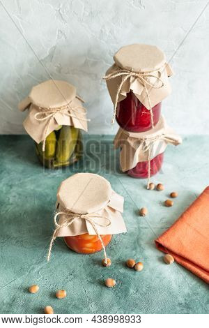 Pickles, Canned Vegetables In Glass Jars And Scattered Nuts And Terracotta Napkin On A Green Texture